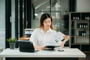 ung skön kvinna skriver på läsplatta och bärbar dator medan Sammanträde på de arbetssätt vit tabell modern kontor foto
