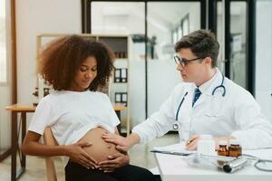 gravid afrikansk kvinna har utnämning med läkare på klinik. manlig gynekolog ob gyn läkare specialist med stetoskop lyssnar till bebis hjärtslag i mors mage. foto