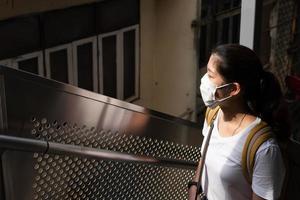ung vacker asiatisk kvinna som bär ansiktsmasker är en social distanseringsriktlinje. hon använder rulltrappan för att gå uppför trappan till himmelstationen. ny normal livsstil, covid-19, coronavirus koncept foto