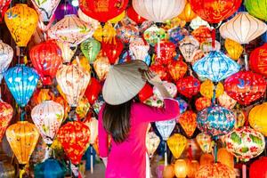 ung kvinna turist i vietnamese traditionell klänning ser på papper dekorativ lyktor i hoi ett gammal stad foto