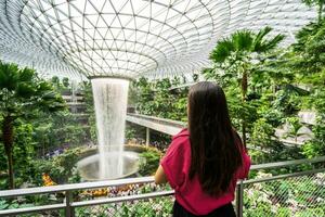 ung kvinna turist ser på de inomhus- vattenfall i changi flygplats, singapore foto