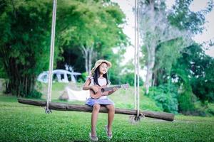 liten asiatisk tjej sitter på trä gunga spelar ukulele medan camping i naturparken foto