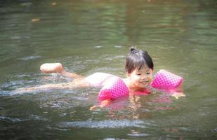 liten asiatisk tjej som bär uppblåsbara ärmar som simmar i naturvatten på semester foto