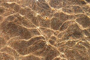 transparent klar vatten hav yta textur med krusningar över sandig strand med sten. abstrakt sommar bakgrund. kopia Plats foto