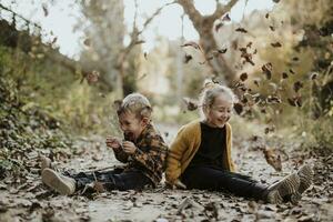 glad flicka och pojke spelar med torr fallen blad medan Sammanträde på gångstig i skog foto