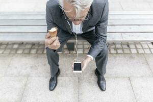 senior affärsman med is grädde kon Sammanträde på bänk ser på cell telefon foto