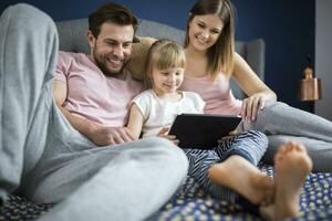 Lycklig familj Sammanträde på säng, tittar på något på digital läsplatta med liten dotter foto