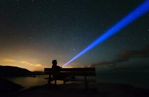 spanien, ortigueira, loiba, silhuett av en man Sammanträde på bänk under starry himmel med blå stråle foto