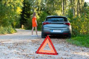 stressande ung kvinna förare liftar och stoppar bilar, ber om hjälp som har problem med brocken bil, använder rött triangel tecken för att varna förare om stopp foto