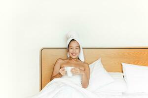 kvinna liggande i säng innehav kaffe råna med en handduk på henne huvud. foto