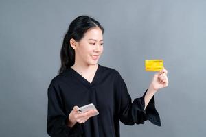 porträtt av en glad ung asiatisk tjej som visar plastkreditkort medan du håller mobiltelefonen på grå bakgrund