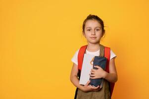 elementärt ålder barn, primär skola studerande flicka bärande ryggsäck, ler sött ser på kamera, orange bakgrund foto