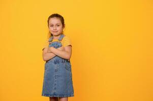 skön liten flicka i blå denim overall, ler ser på kamera, Framställ med vapen vikta över orange bakgrund foto