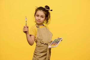 äkta barn flicka bär en beige förkläde och frisyr med pensel, innehav palett kniv och färgrik akvareller foto