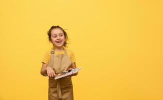 busig unge flicka i beige förkläde, primär skola studerande doppning paintbrush in i vattenfärg målar, ler på kamera foto