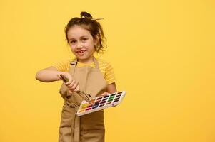 förtjusande liten flicka i beige förkläde, primär skola studerande doppning paintbrush in i vattenfärg målar, ler på kamera foto