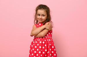 underbar blygt 4 år unge i prickar klänning kramar själv, ljuv ler Framställ på rosa vägg bakgrund med Plats för text. självsäker porträtt av skön bebis flicka för reklam foto