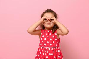 leende förskola flicka i en rosa polka punkt klänning, imiterar en binokulär med henne händer och ser genom Det. förtjusande barn uttrycker positiv känslor Framställ på rosa bakgrund foto