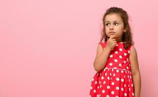 en söt förtjusande flicka av förskola ålder, 3-4 år gammal, utseende eftertänksamt till de sida , innehav henne finger på henne haka, Framställ på en rosa bakgrund med kopia Plats foto