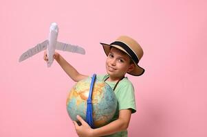 leende skola pojke ser på kamera medan spelar med papper flygplan och en klot, stående isolerat över rosa bakgrund med kopia Plats. turism, resa, geografi kunskap begrepp foto