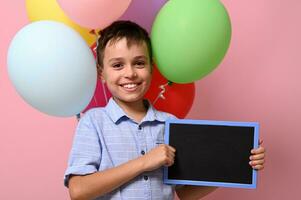 förtjusande leende skol med tömma tom svarta tavlan i hans hand stående mot flerfärgad ballonger på rosa bakgrund med kopia Plats foto