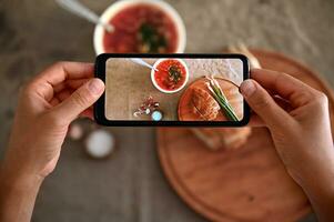 topp se av händer innehav smartphone och fotografering en skål med nyligen kokta ukrainska traditionell soppa- borscht med färsk grön rå lök . mobil slipa i leva se läge foto