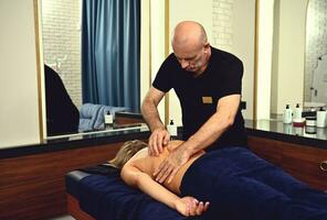 professionell massör masse kvinnas tillbaka i en wellness klinik. en mogna kiropraktor ger en terapeutisk massage till en klient av de spa Centrum. foto