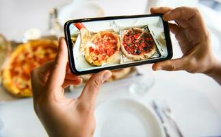händer innehav smartphone och tar bild av utsökt nyligen bakad pizza eras på en tabell med vit bordsduk. mobil telefon i leva läge regimen foto
