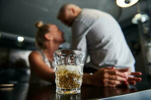 fokus på en glas av alkoholhaltig dryck och is kuber mot en suddig bakgrund av en mitten åldrig europeisk heterosexuell par flirta tillsammans på en bar. foto