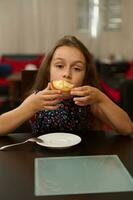 äkta söt liten barn flicka Sammanträde på tabell och njuter äter färsk ljuv sugary efterrätt - en tartlet med citron- foto