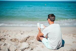 ung man Sammanträde på de strand läsning bok foto