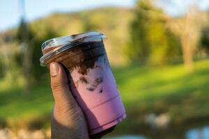 en hand innehav en glas av choklad på topp av rosa mjölk med is i en plast kopp. foto