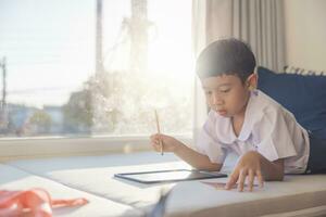 Lycklig asiatisk pojke 6-7 år gammal, svart hår, vit hud, bär vit skjorta och blå shorts skrivning på en svarta tavlan. barn i skola uniformer av thailand. begrepp handla om utbildning, spela, kreativitet. foto