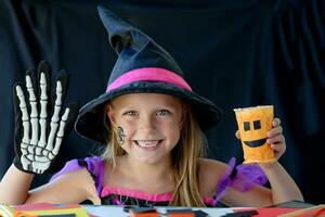 en liten flicka i en häxa kostym utseende på de kamera och skrattar, innehav en glas pumpa och en skelett handske i henne händer på svart bakgrund. foto