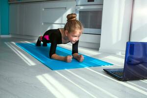 liten flicka håller på med yoga planka och tittar på uppkopplad lektioner på bärbar dator, utövar i de kök. foto