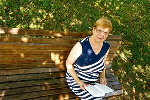 senior kvinna läsning en bok i de parkera på en solig dag foto