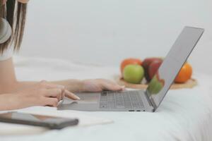 effektiv ung tusenåriga flicka Sammanträde på en säng i de morgon, användningar bärbar dator dator och äter croissanter och drycker kaffe för frukost foto
