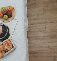 effektiv ung tusenåriga flicka Sammanträde på en säng i de morgon, användningar bärbar dator dator och äter croissanter och drycker kaffe för frukost foto