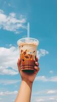 hand innehav iced kaffe i en plast kopp med en blå himmel och moln bakgrund. ai genererad foto