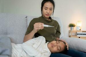 asiatisk kvinna ta vård av liten sjuk dotter. sjuk barn liggande på säng under filt, med orolig. enda mamma tar vård av sjuk dotter på Hem. barn har en hög feber. omslag på de soffa och sjuk foto