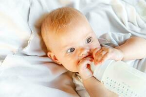 söt liten nyfödd flicka dricka mjölk från flaska och ser på kamera på vit bakgrund. spädbarn bebis sugande äter mjölk näring liggande ner på spjälsäng säng på Hem. moderskap Lycklig barn begrepp. foto
