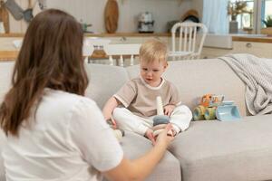 Lycklig familj på Hem. mor och bebis pojke spelar med leksaker i soffa på Hem inomhus. liten litet barn barn och barnvakt barnskötare har roligt tillsammans. ung kvinna mamma unge son resten i levande rum. foto