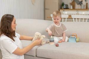 Lycklig familj på Hem. mor och bebis pojke spelar med leksaker i soffa på Hem inomhus. liten litet barn barn och barnvakt barnskötare har roligt tillsammans. ung kvinna mamma unge son resten i levande rum. foto