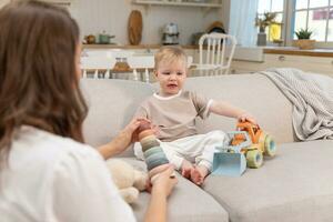 Lycklig familj på Hem. mor och bebis pojke spelar med leksaker i soffa på Hem inomhus. liten litet barn barn och barnvakt barnskötare har roligt tillsammans. ung kvinna mamma unge son resten i levande rum. foto