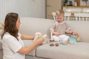 Lycklig familj på Hem. mor och bebis pojke spelar med leksaker i soffa på Hem inomhus. liten litet barn barn och barnvakt barnskötare har roligt tillsammans. ung kvinna mamma unge son resten i levande rum. foto