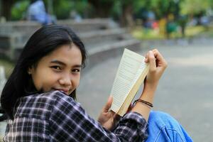 Lycklig ung asiatisk kvinna högskola studerande med leende ansikte njut av läsa en bok för examen i de parkera foto