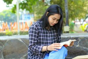 Lycklig uppmärksam ung asiatisk kvinna högskola studerande läsning en bok i de parkera, utbildning begrepp foto