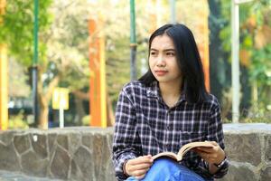 Lycklig ung asiatisk högskola studerande läsa en bok i de parkera ser bort foto