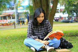 Lycklig och succes kvinna asiatisk högskola studerande njuter läsa en bok i de parkera foto
