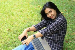 upphetsad skön asiatisk studerande använder sig av bärbar dator och smartphone och läsning bok till studerar och planera henne arbete foto